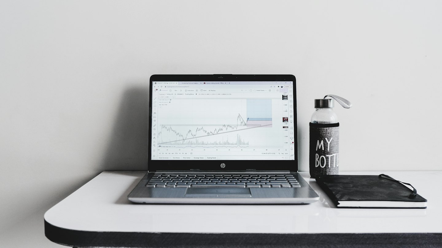 Un laptop con grafici finanziari accanto a un taccuino e una bottiglia d'acqua.